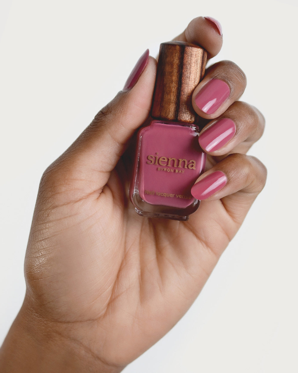 Medium tanned skin hand wearing Heartspace raspberry sorbet nail polish and holding bottle with timber lid by Sienna Byron Bay