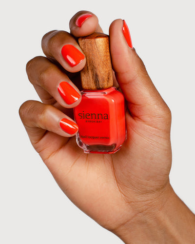 Warm strawberry red nail polish hand swatch on medium skin tone holding sienna bottle