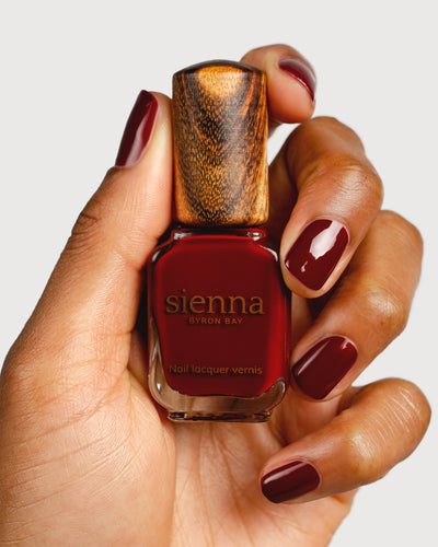 Organic mid-tone red nail polish hand swatch on medium skin tone holding sienna bottle close-up